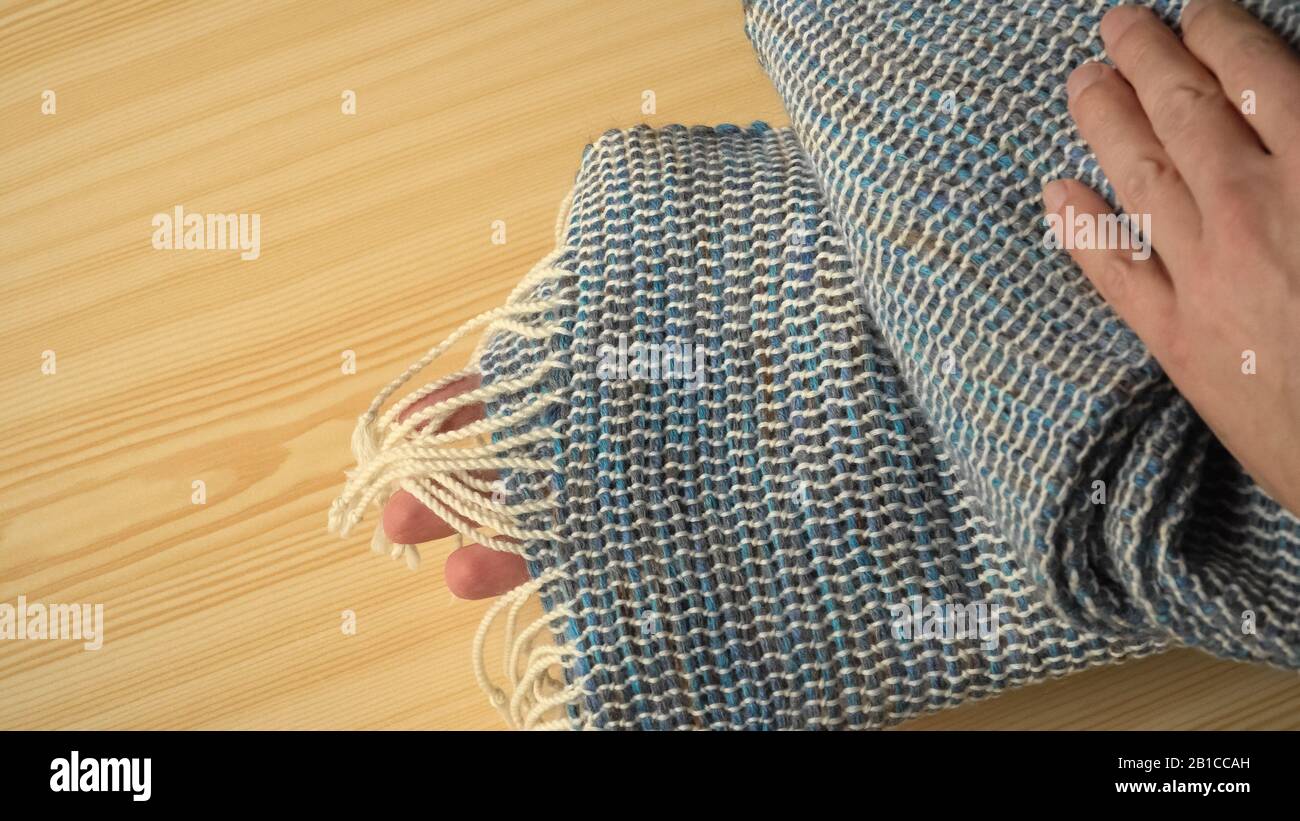 A person demonstrates a handwoven wool scarf made in looms. Woven fabric with white stripes and darker bands in blue-green tones in knitter's hands. Stock Photo