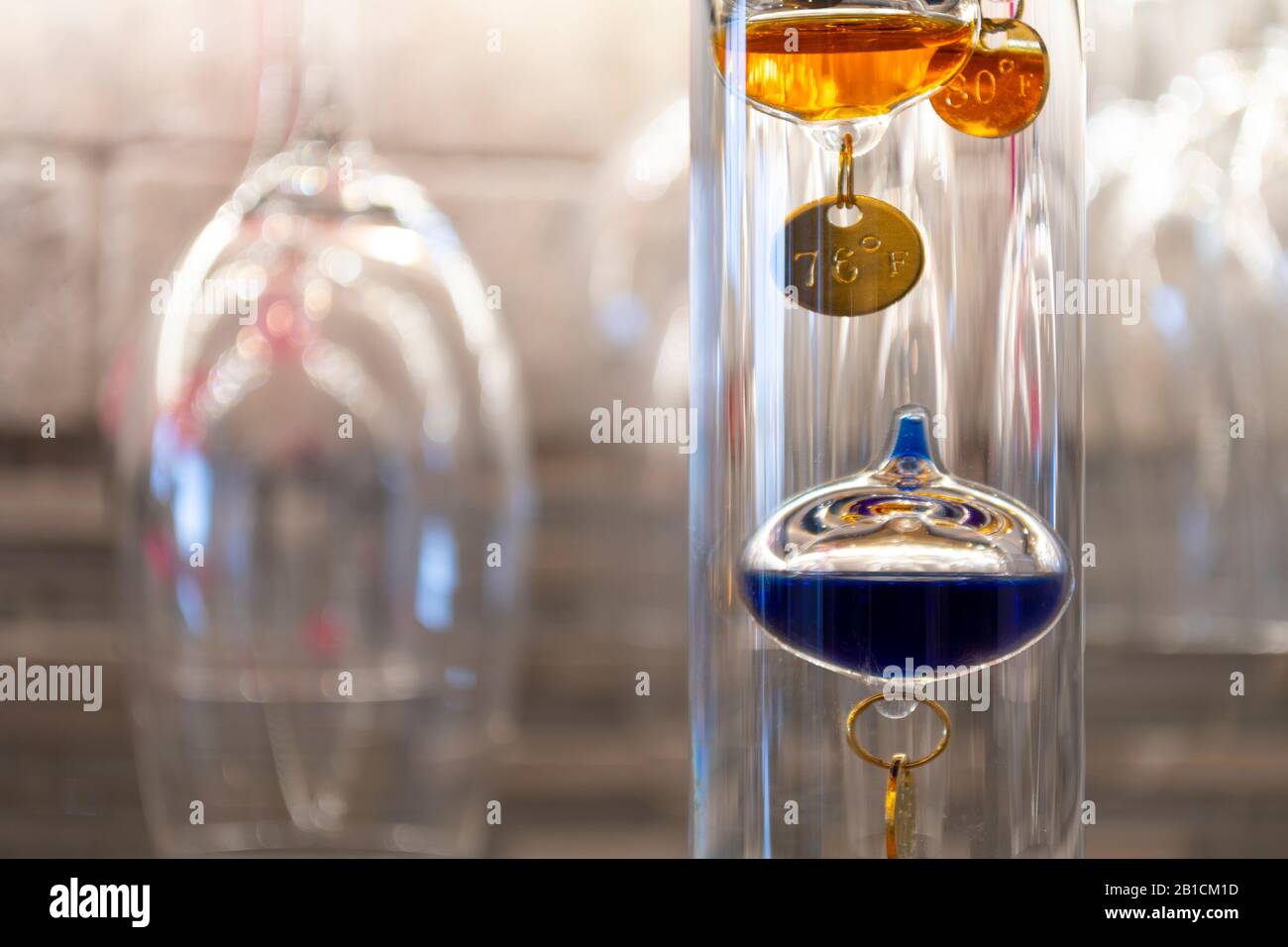 A colorful, glass, liquid filled Galileo Thermometer measures indoor room temperature in a wine cellar with wine glasses hanging behind. Stock Photo