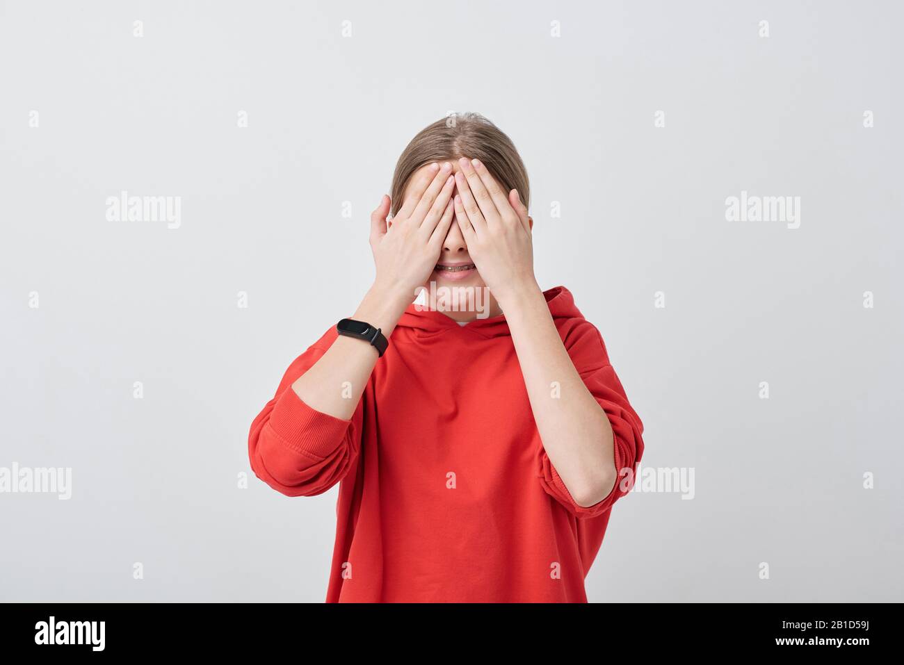 Smiling teenage girl in red sweatshirt covering eyes with hands while playing hide-and-seek Stock Photo