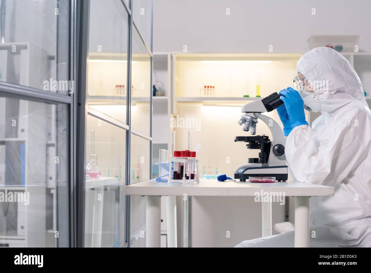 Side view of medical scientist in protective mask using microscope while studying dangerous coronavirus in laboratory Stock Photo