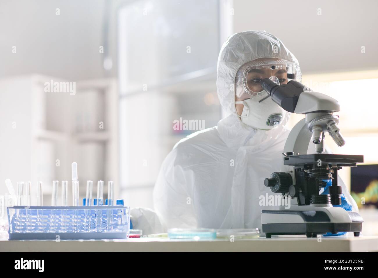 Concentrated Asian virology researcher in protective suit using microscope while testing virus sample in laboratory Stock Photo