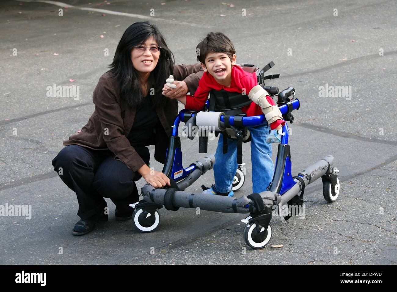 Asian mother  helping little disabled son  walk in his walker outdoors  on sidewalk or pavement Stock Photo