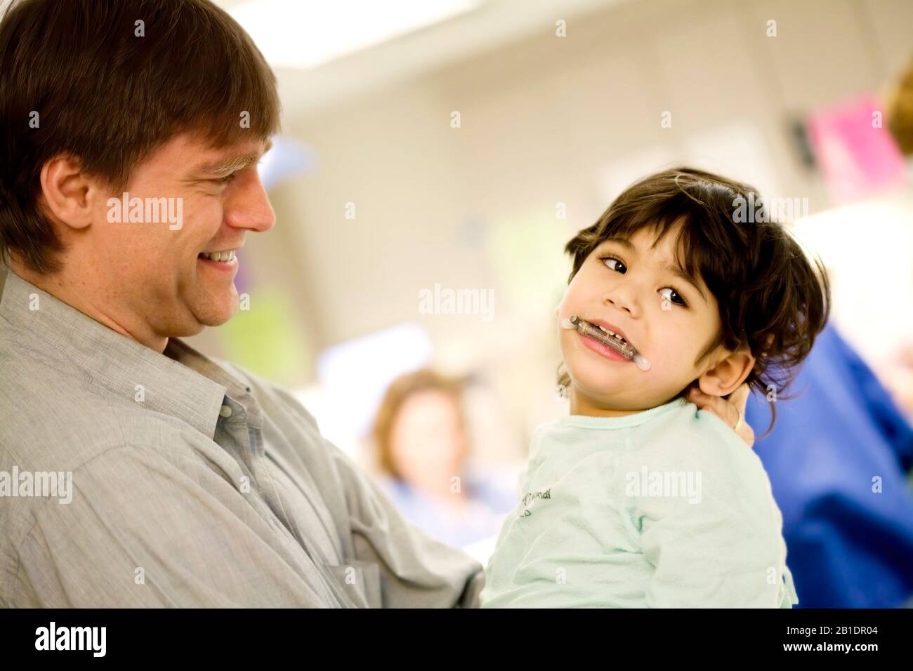 Caucasian father holding sick disabled little boy in surgical gown in hospital before surgery. Child has medical mouth device in place. Stock Photo