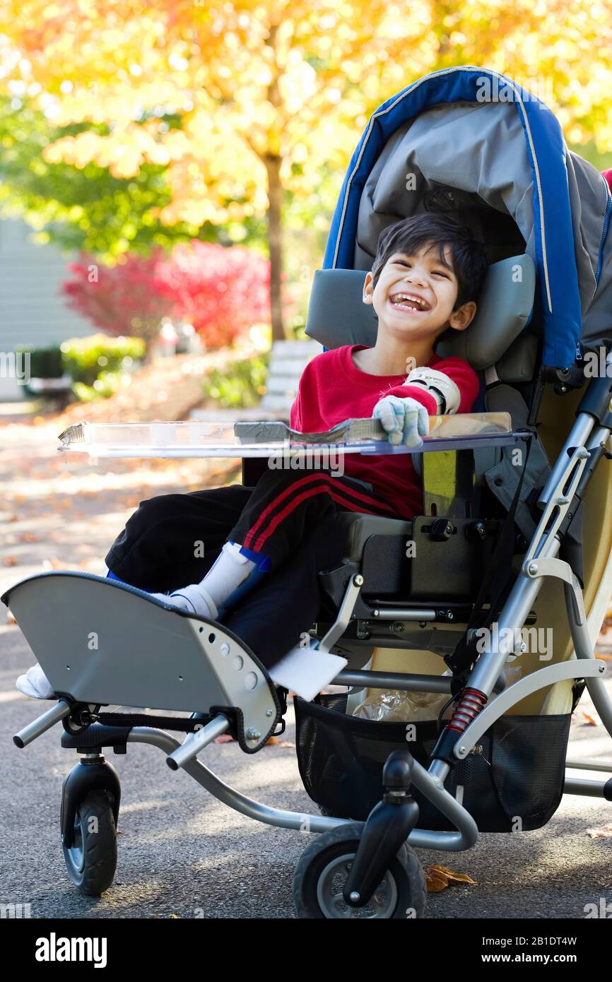 Handsome little disabled child sitting in wheelchair at park on sunny day outdoors with colorful yellow leaves on trees in background Stock Photo