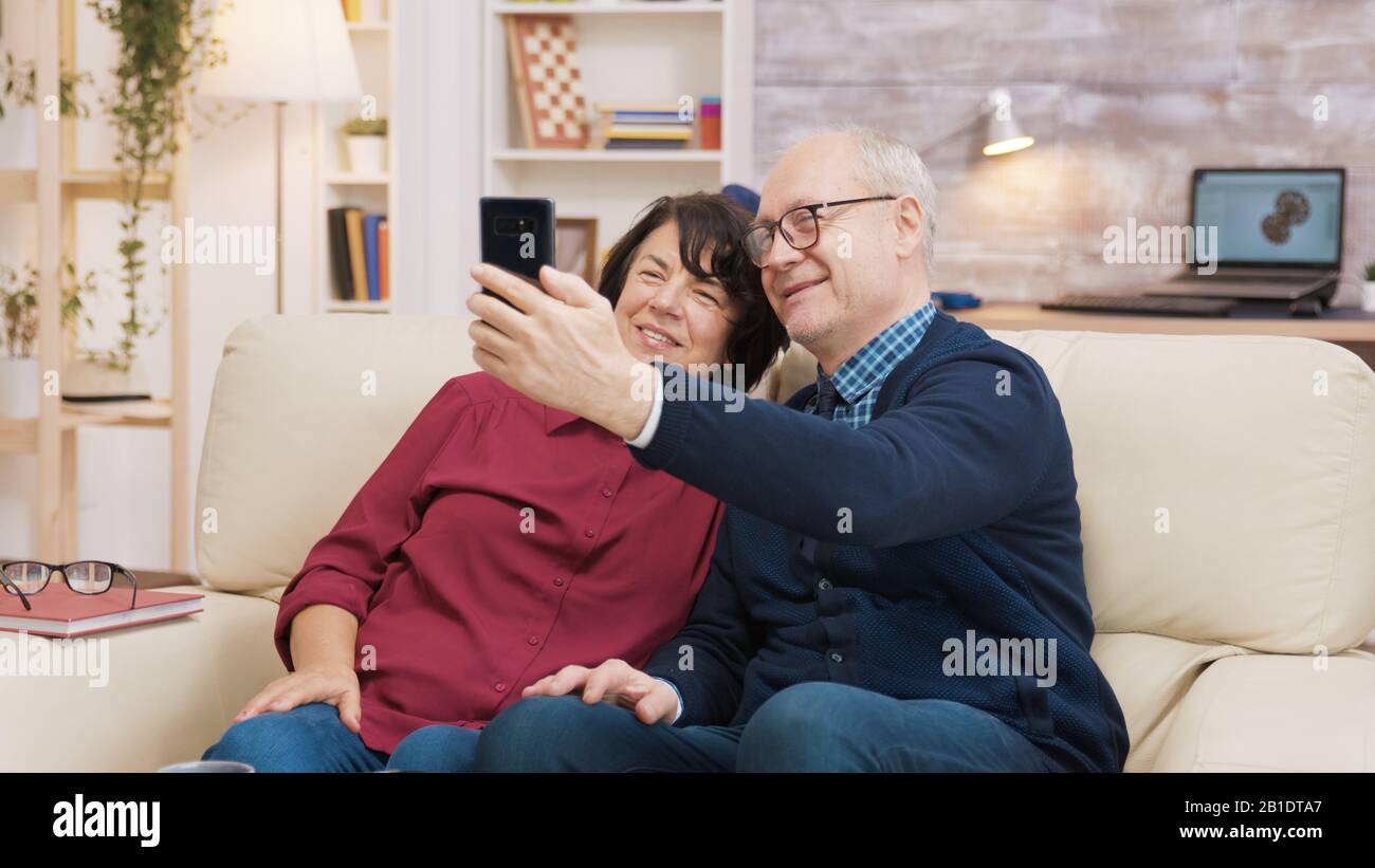 Happy senior couple sitting on sofa taking a selfie in the living room ...