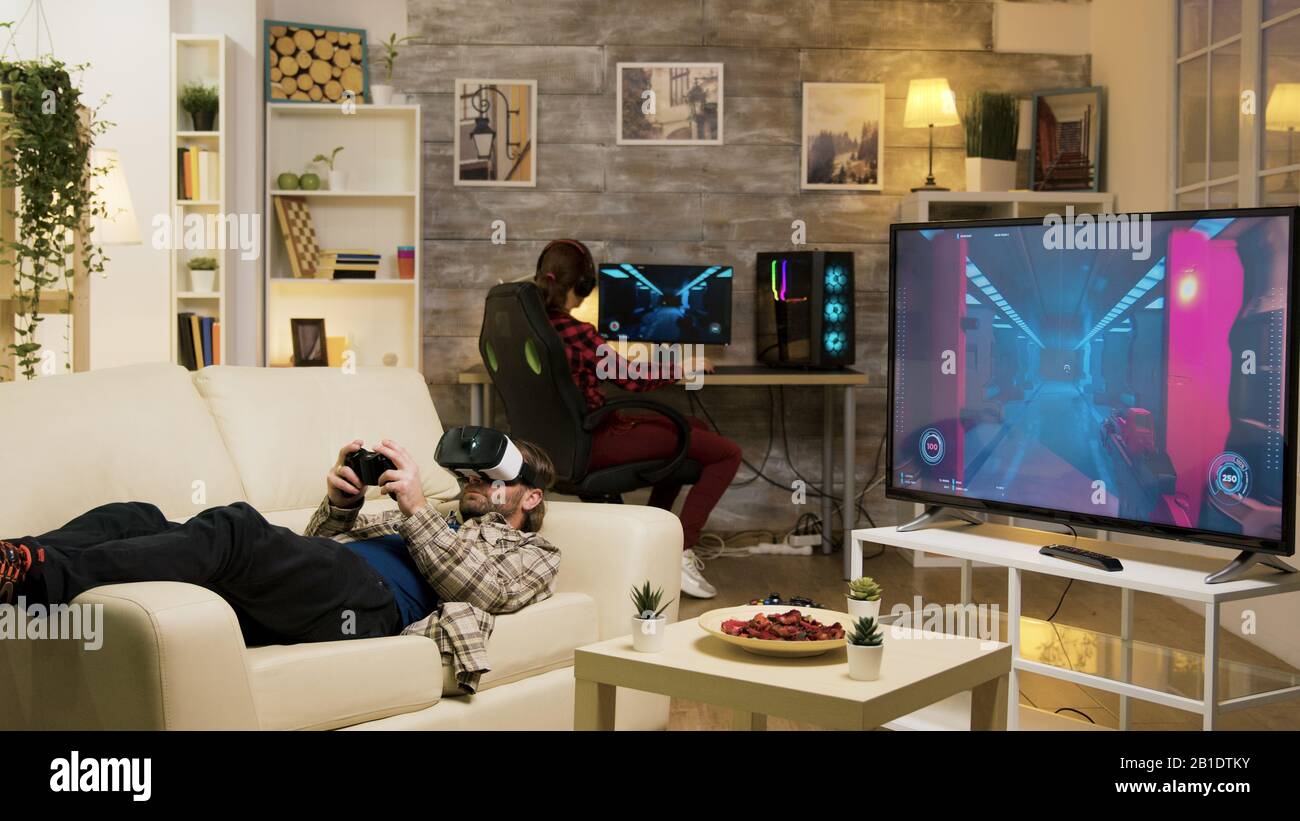 Man lying on sofa playing video games using vr headset with his girlfriend in the background playing on computer. Stock Photo