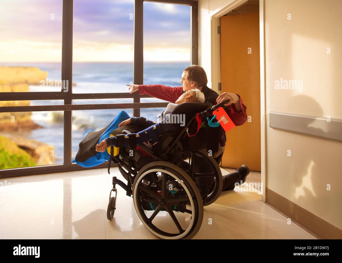Father kneeling next to disabled son in wheelchair looking out hospital window at ocean and cliffs outdoors. Child recently had head surgery. Stock Photo