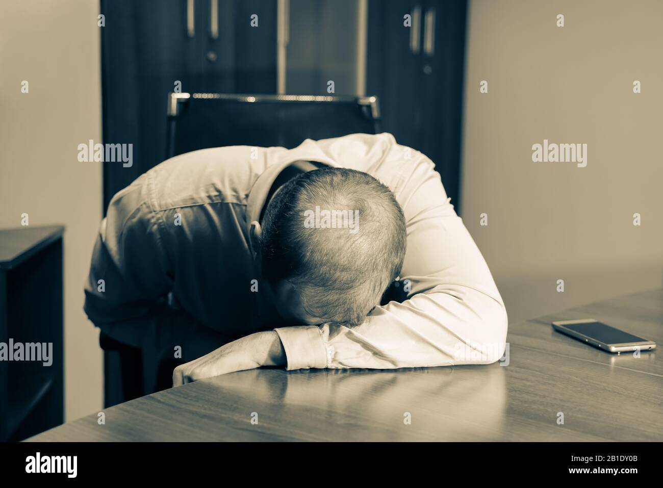 Middle aged man is at the desk in the office, depressed and exhausted. He is just sitting at the working place with his head on the table. Sepia color Stock Photo