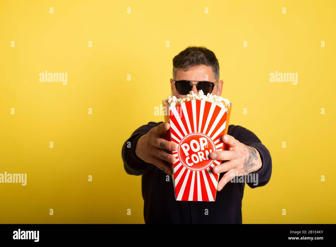 Man with defocused sunglasses showing a box full of popcorn over yellow background. Stock Photo