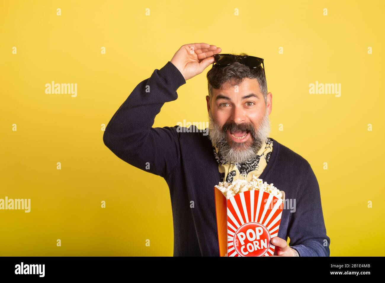 Man with white beard deeply entertained by raising sunglasses eating popcorn on yellow background. Stock Photo