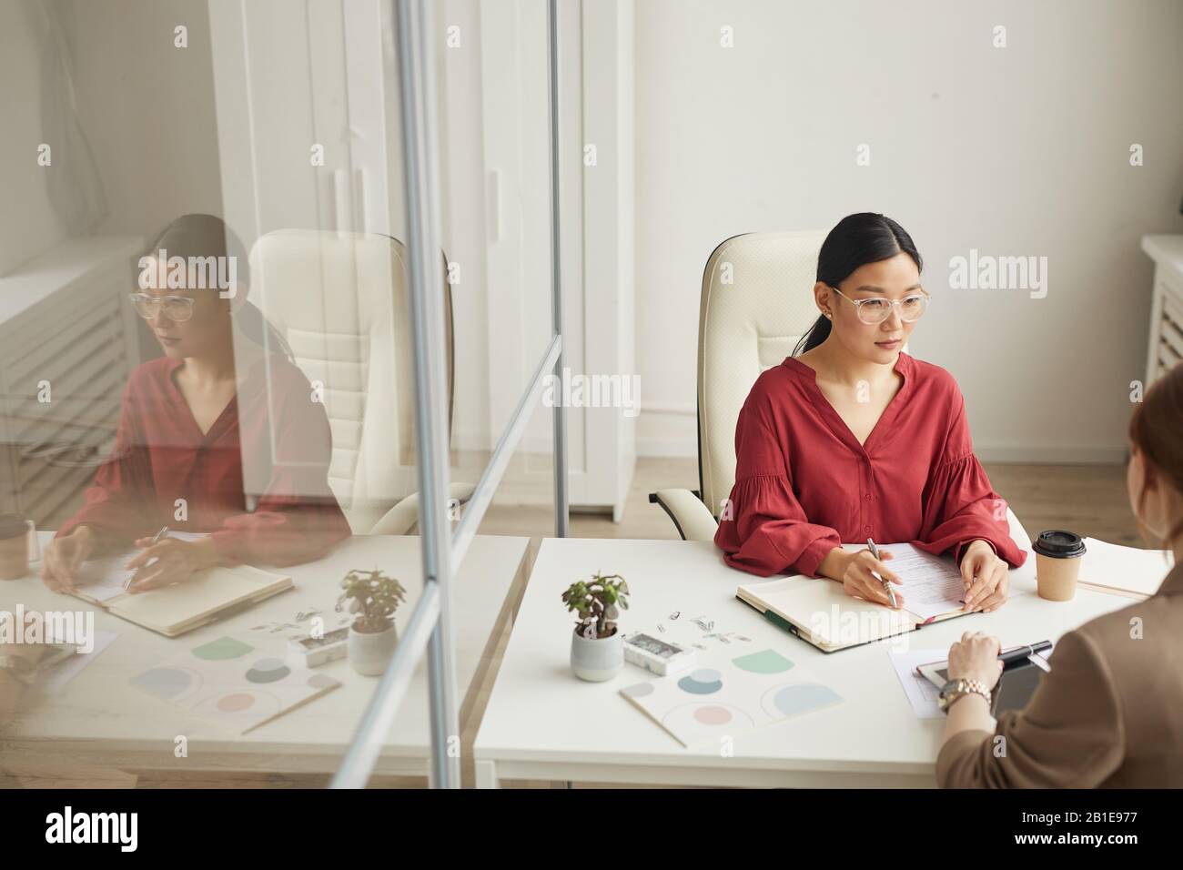 High angle view at modern Asian businesswoman talking to client while working at desk in white office cubicle, copy space Stock Photo