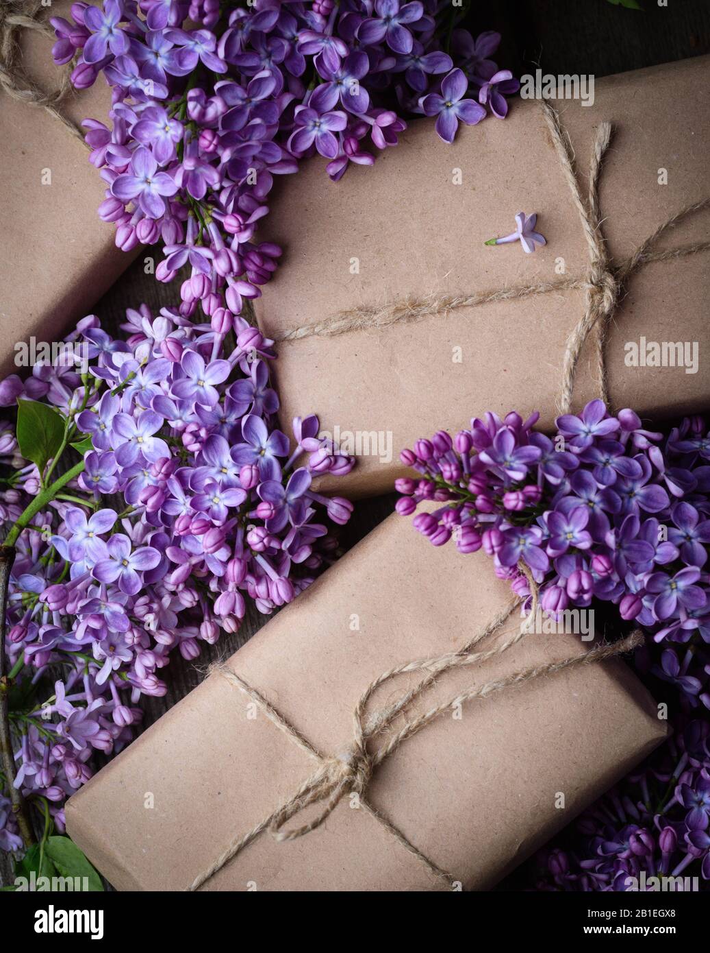 Handmade gift boxes wrapped with craft brown paper, decorated with fresh purple lilac flowers on a vintage wooden surface. Spring holidays background. Stock Photo