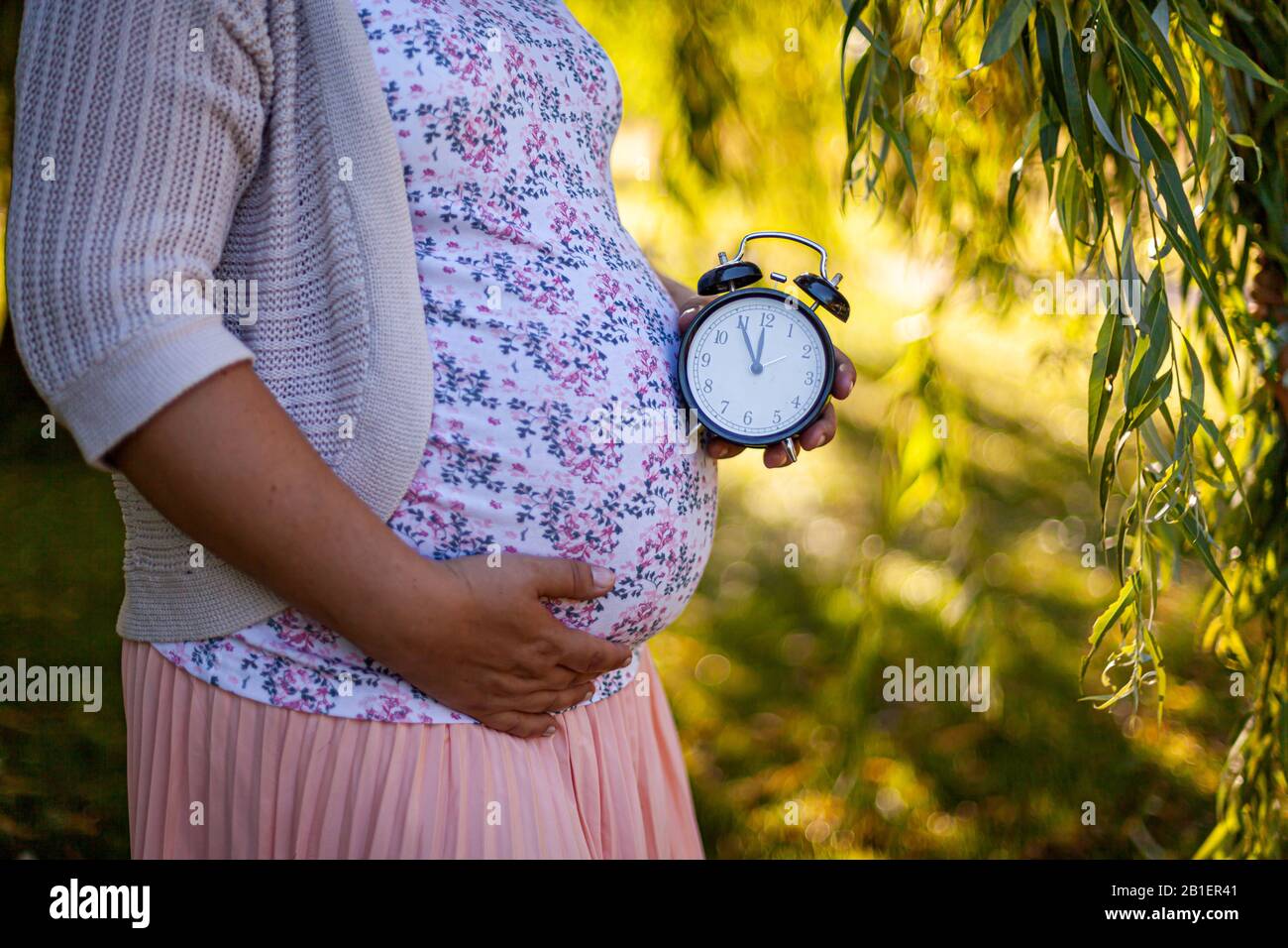A pregnant women in a white dress in park holding a clock Stock Photo