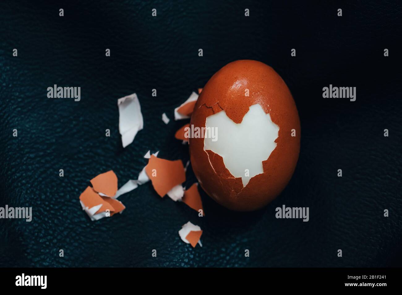 Cracked hard boiled brown egg with heart shaped shell on black leather background. Minimal Easter holiday concept. Stock Photo