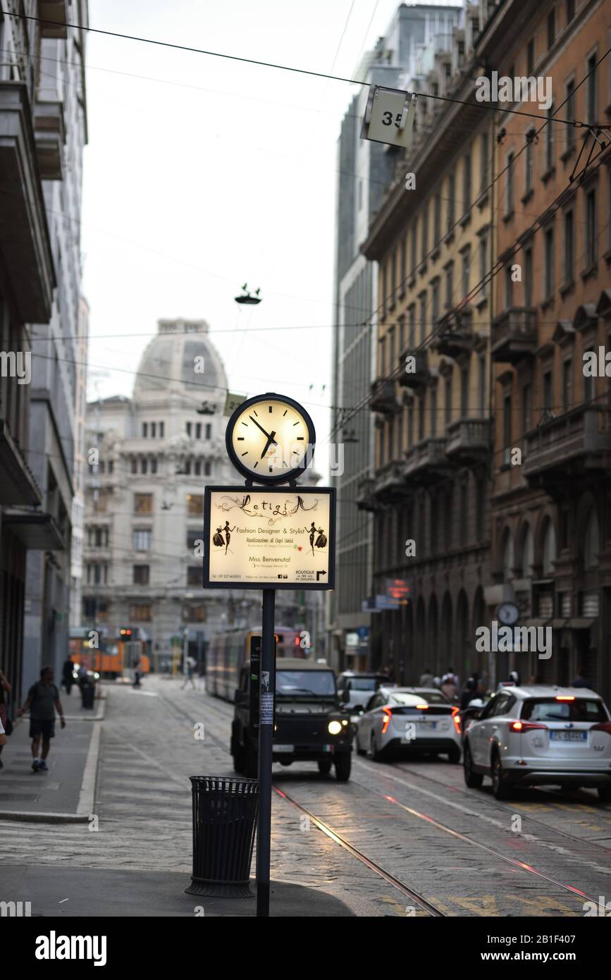 An illuminated street clock and advertisement - street furniture, traffic on Via Giuseppe Mazzini with Palazzo Meroni at dusk, Milan, Italy Stock Photo