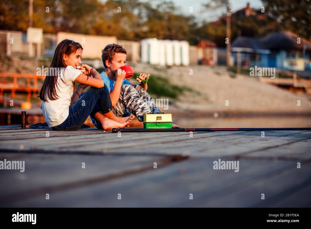 Two young cute little friends, boy and girl talking, drinking tea, eating sandwiches and fishing on a lake in a sunny summer day. Kids are playing. Fr Stock Photo