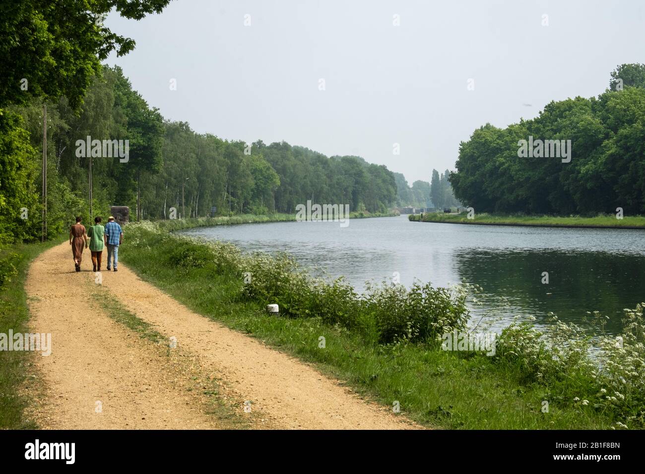 People are walking on a path along the Bocholt-Herentals canal in Mol. This peaceful area of the canal is bordered by trees. Stock Photo
