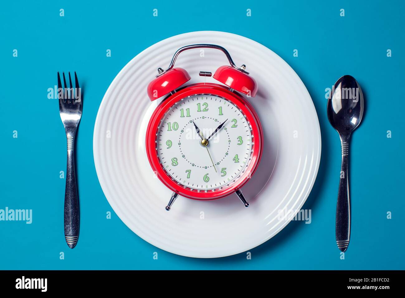 Red alarm clock on the white plate with spoon and fork on the color background. Food and diet concept Stock Photo