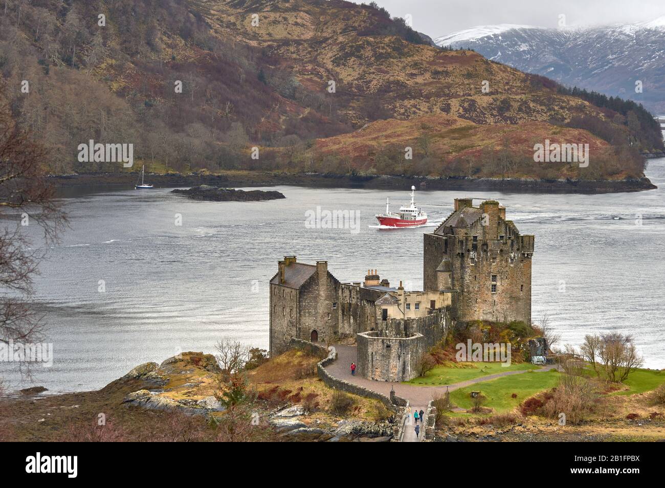 EILEAN DONAN CASTLE LOCH DUICH DORNIE SCOTLAND WINTER THE CASTLE OVERLOOKING THE WATERS OF THE LOCH WITH RED BOAT AND PEOPLE ON THE BRIDGE Stock Photo