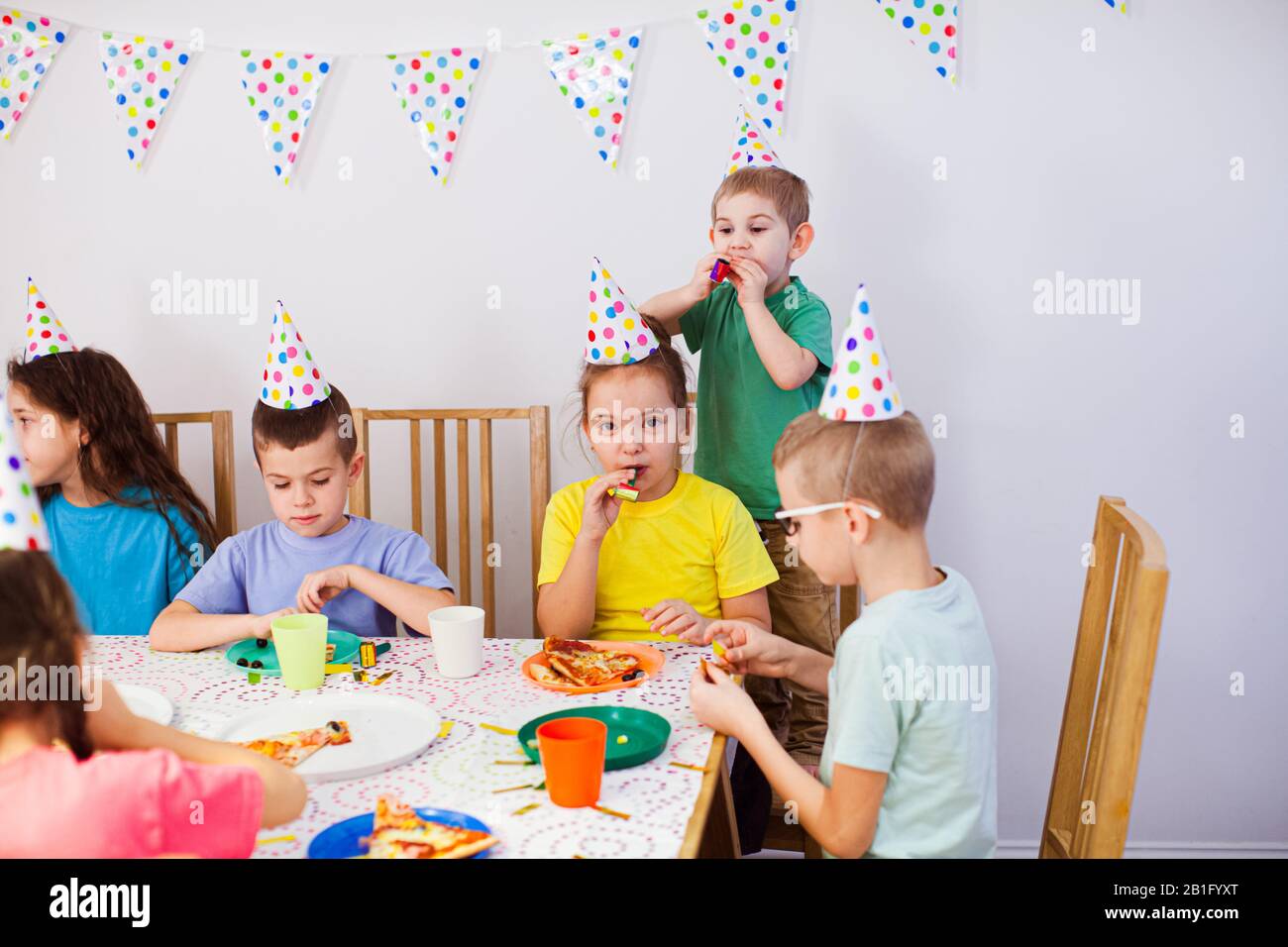 Joyful kids eating pizza at big table. Children wear birthday hats