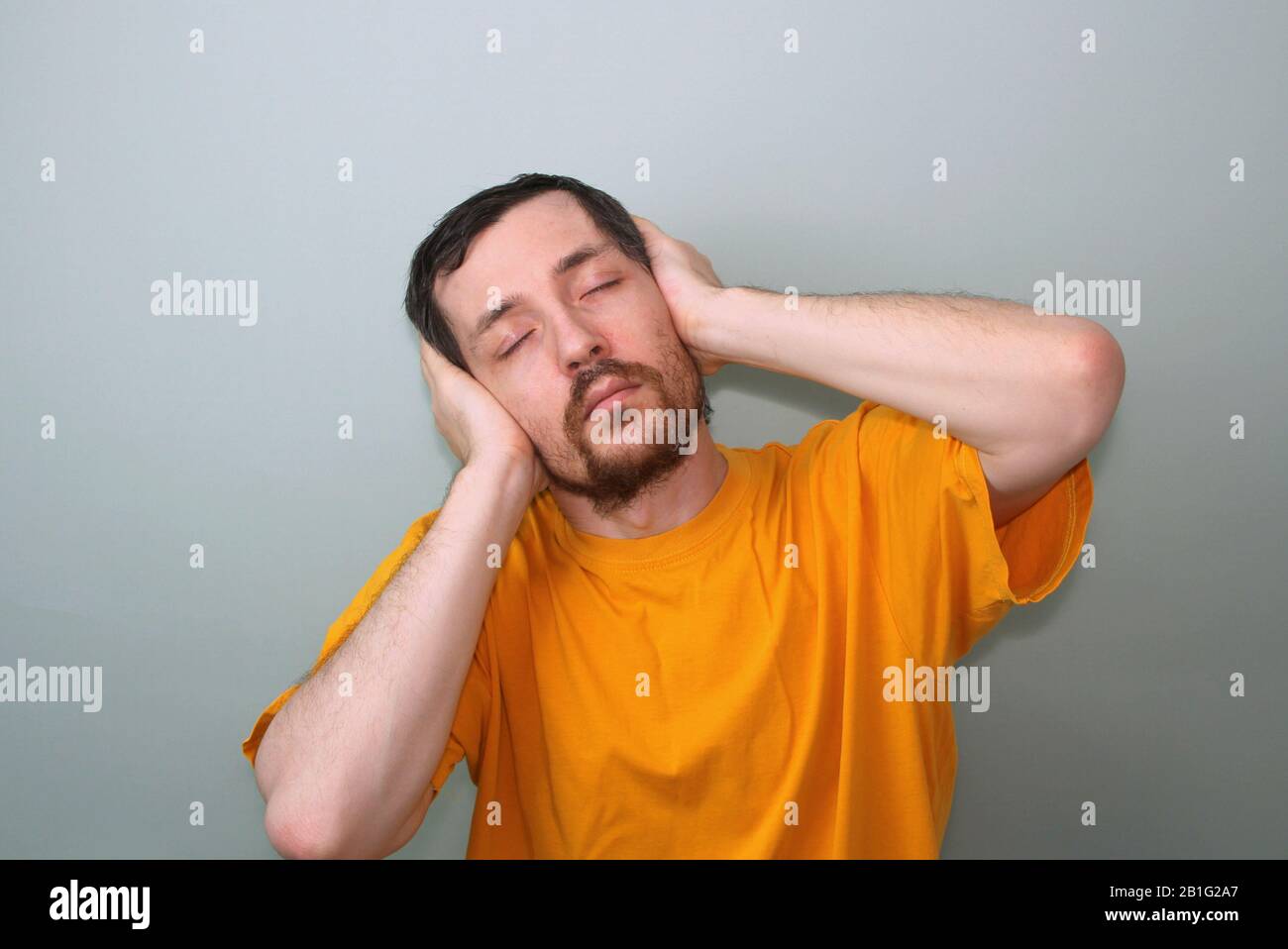 Middle-aged man with a mustache and a brunette beard in a yellow T-shirt on a gray background with closed eyes he covered his ears with the palms of his hands and bent as if he was listening to music. Stock Photo
