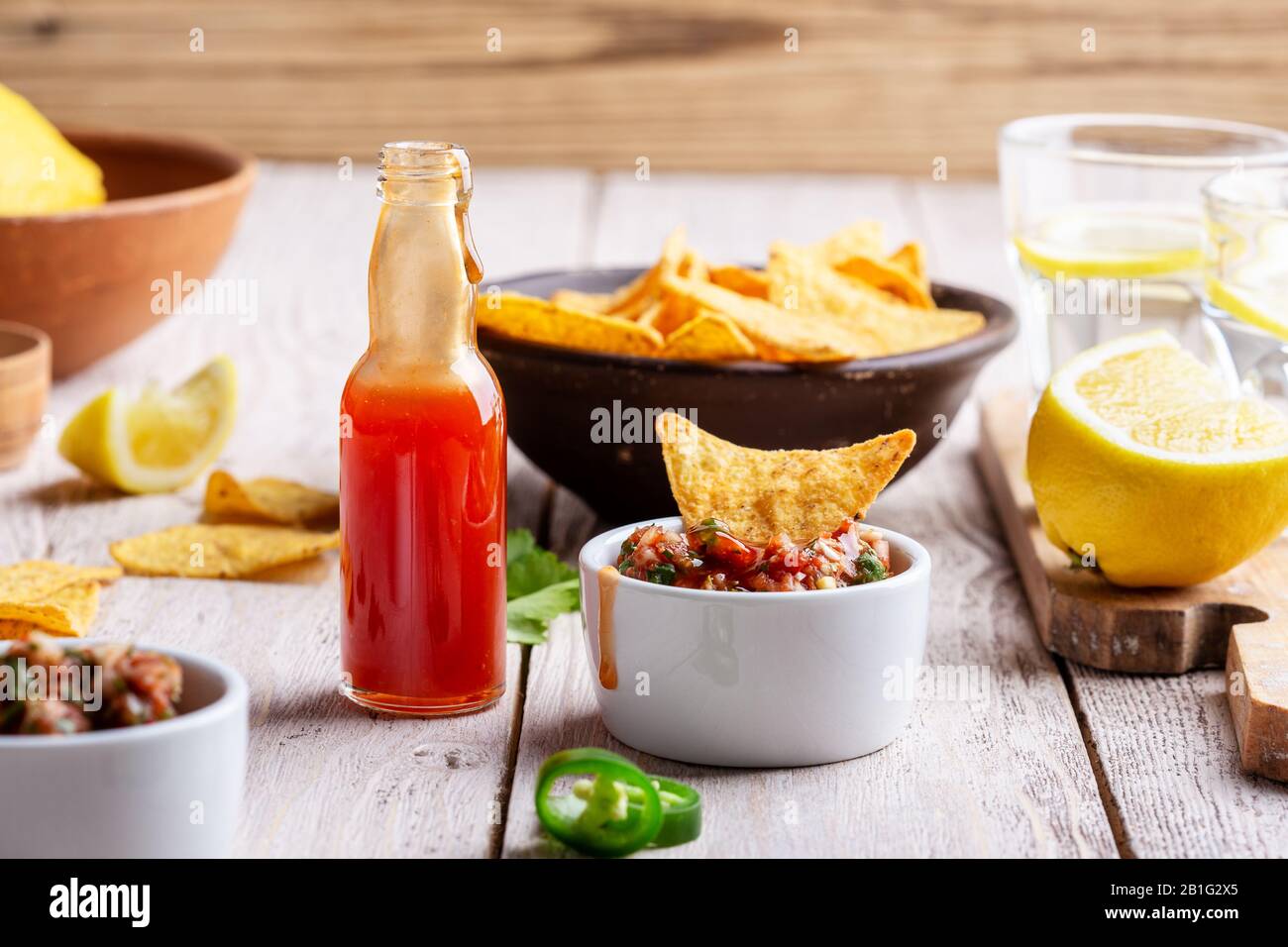 Two bowls full of salsa dip, hot red chili sauce bottle and tortilla chips on rustic wooden table, sause being added to one of the bowls Stock Photo