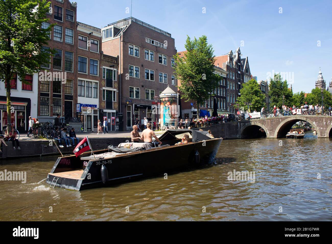 View of people riding a open boat on canal doing a cruise tour in Amsterdam. Many people hang out on street and crossing Armbrug bridge. It is a sunny Stock Photo