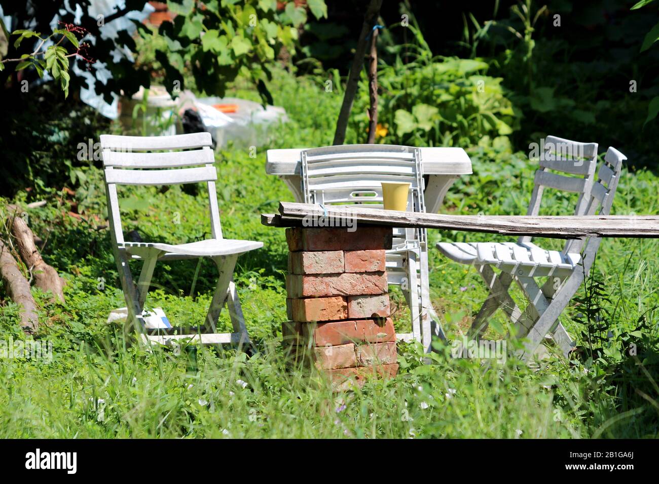 White old vintage retro wooden garden furniture next to improvised table supported by red bricks surrounded with uncut grass and garden plants Stock Photo