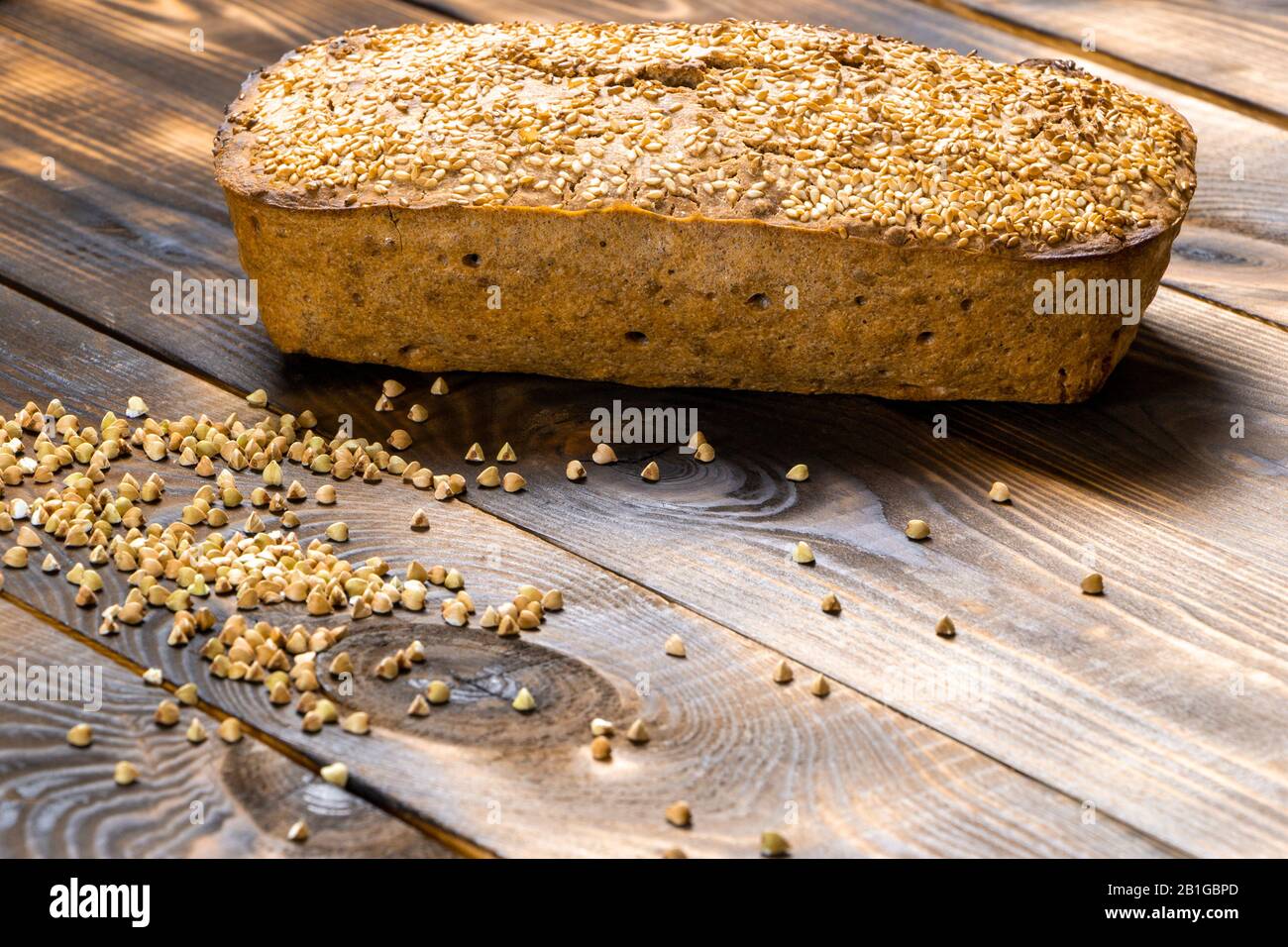 Gluten-free buckwheat bread with a golden brown crust, sprinkled with sesame seeds, lies on a wooden table. Healthy homemade recipe. Grains of green Stock Photo