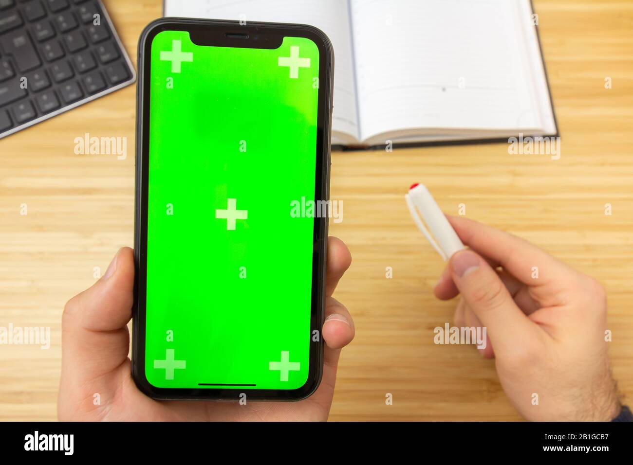 Mockup image of hand holding black mobile phone with green screen and red liner on the office table on the background with stationery tools. San Stock Photo