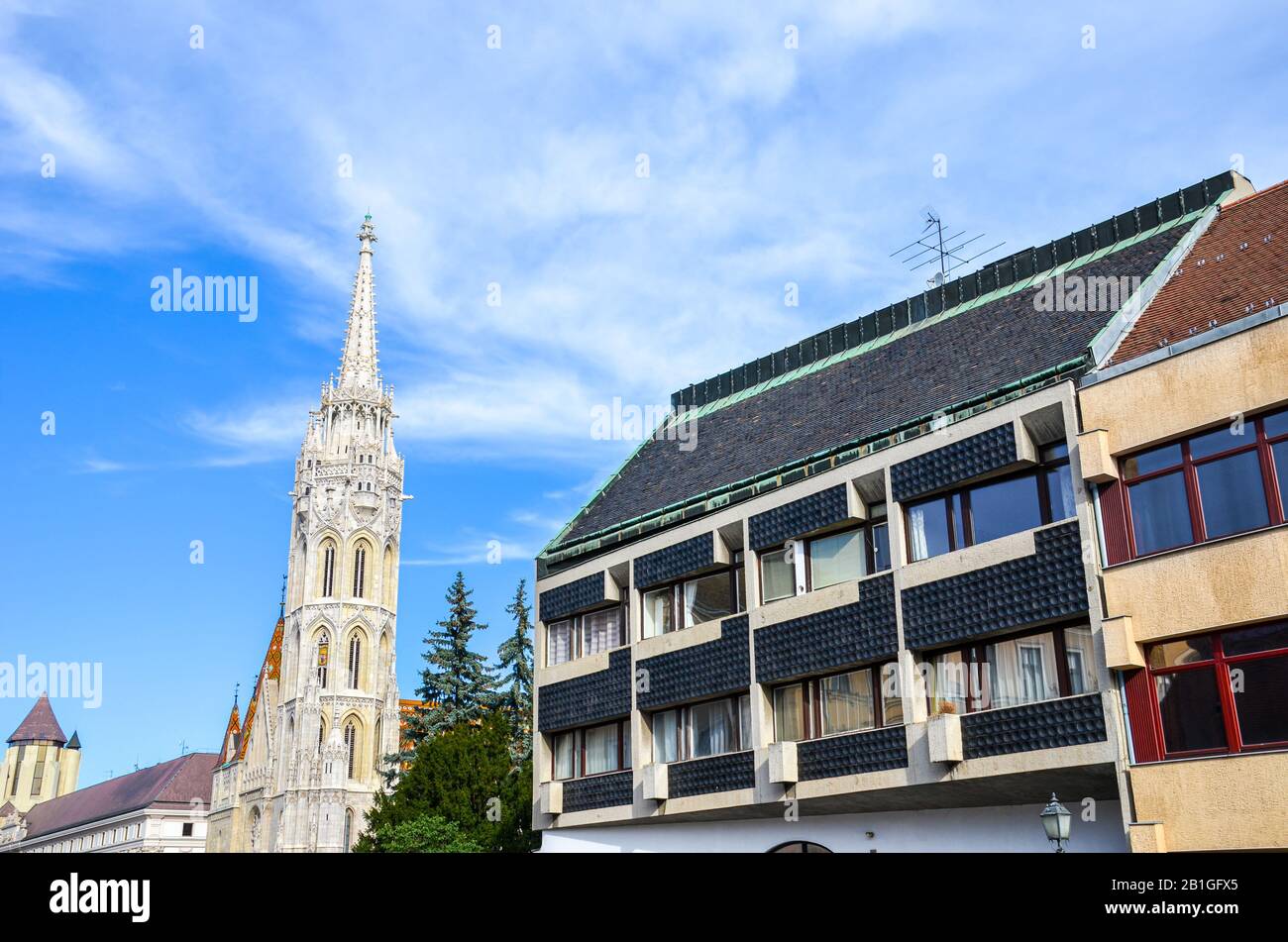 Spire of the Matthias Church in Budapest, Hungary on a horizontal photo with adjacent Socialist building. Roman Catholic church built in the Gothic style. Blue sky and white clouds. Eastern Europe. Stock Photo