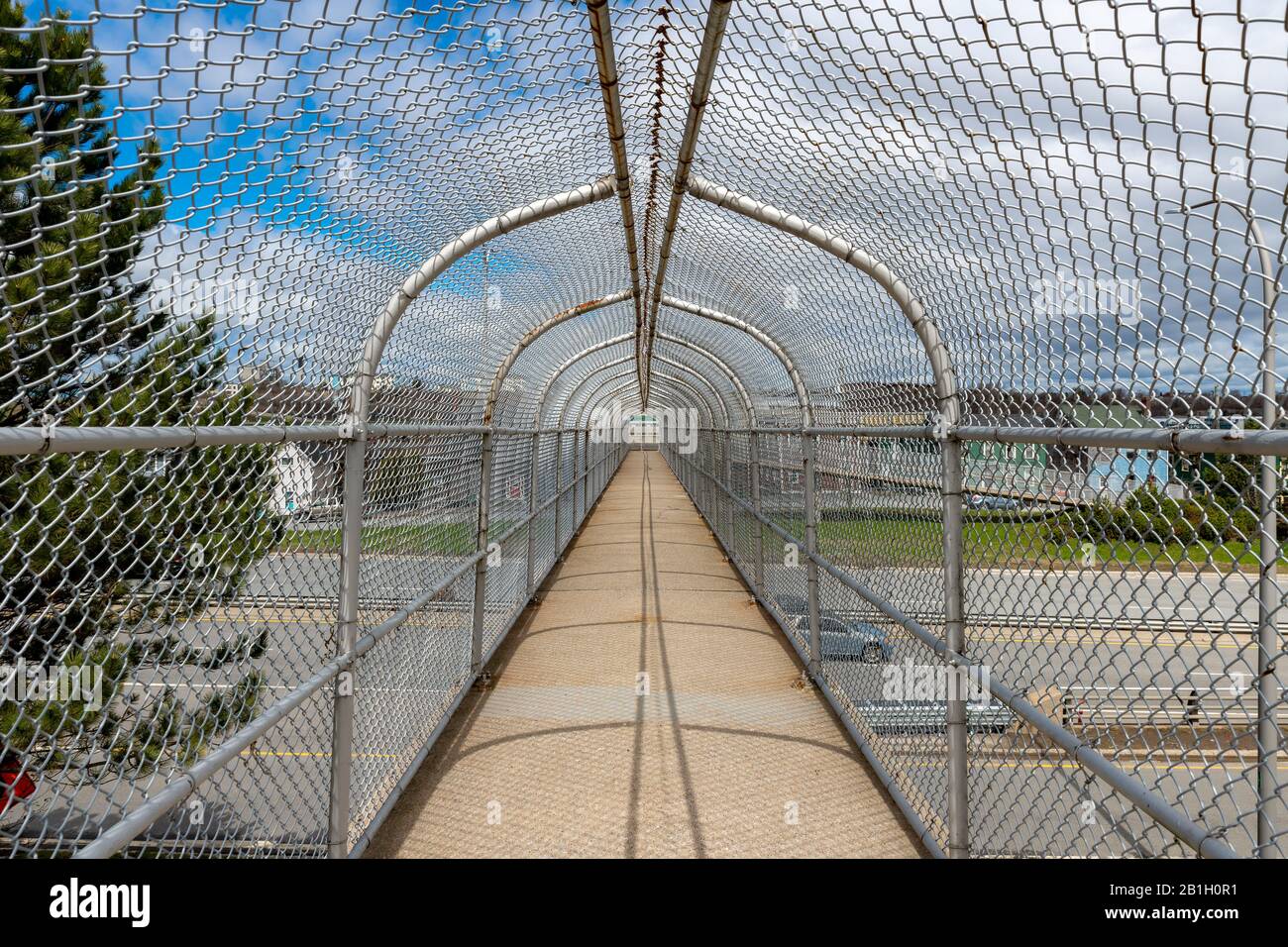 Inside a fenced in walkway over a highway. The walkway has a concrete floor and chain link fencing enclosing the sides and top.Blue and cloudy sky. Stock Photo