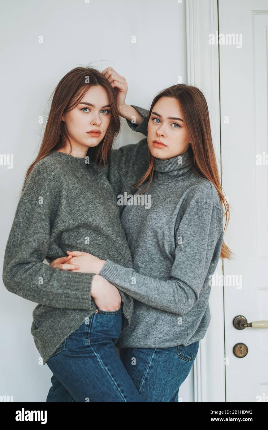Young beautiful long hair girls sisters twins in casual grey sweaters and jeans on background of white door Stock Photo