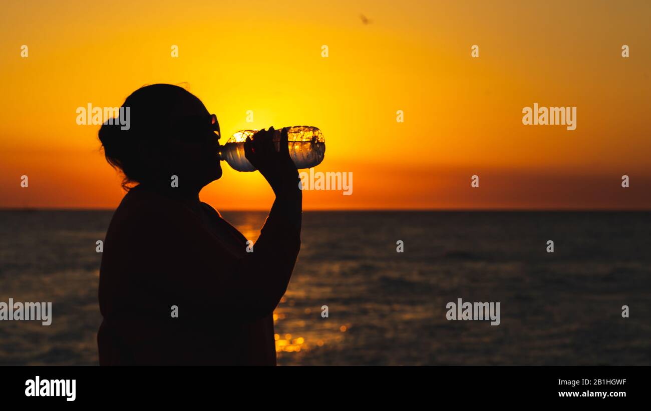 Silhouette of a girl drinking water near the beach whit a orange sky at sunset Stock Photo