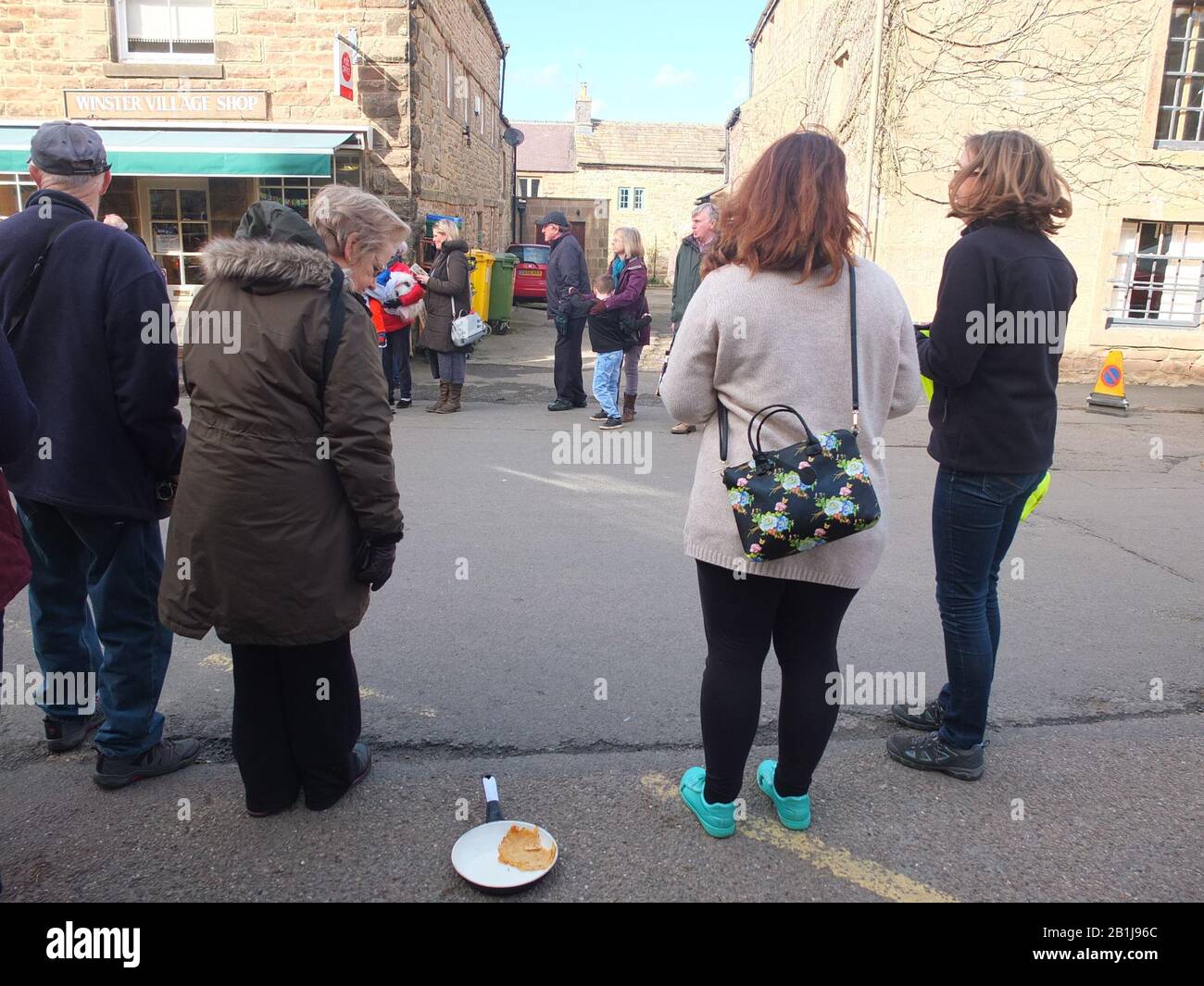 Spectators on village Main Street during Winster Pancake Races lady looks at abandoned frying pan on floor Shrove Tuesday Pancake Day Derbyshire Stock Photo