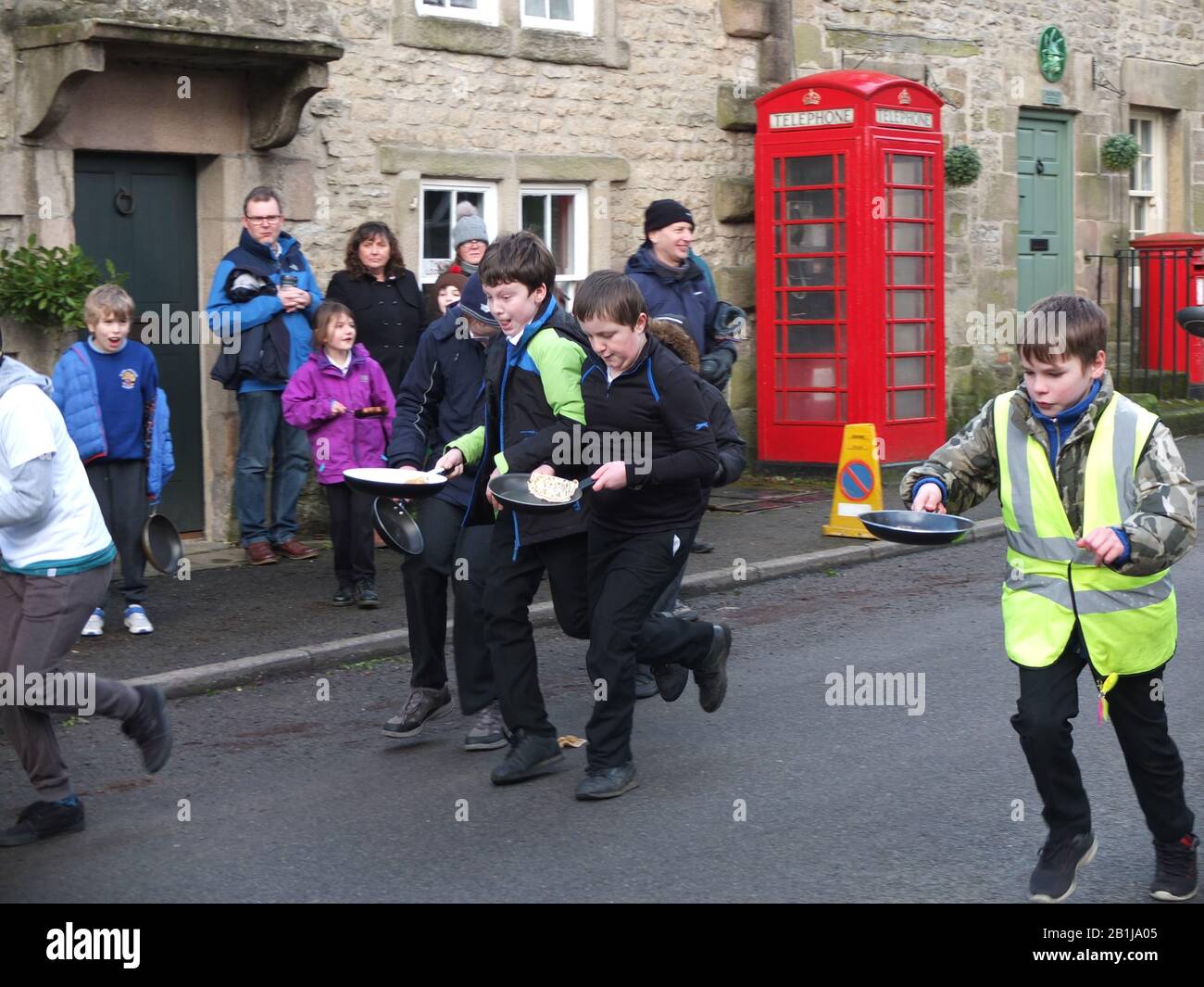 Schoolchildren race down village main street with frying pans and pancakes during Winster Pancake Races Shrove Tuesday Pancake Day Derbyshire UK Stock Photo