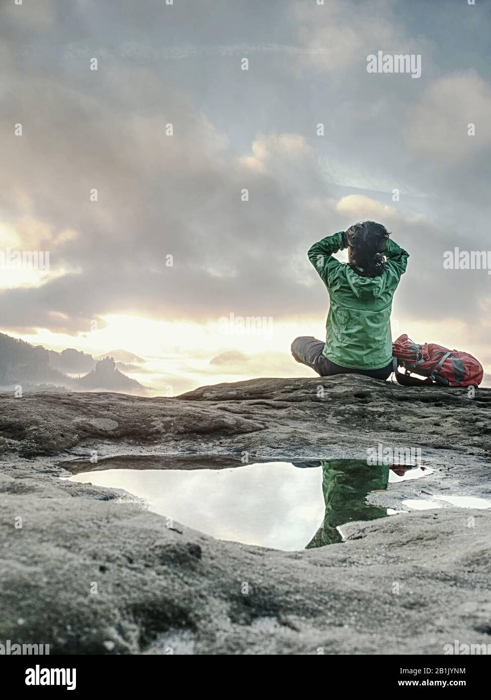 Beautiful girl in a hat and green jacket meditate while sitting on a stone in the mountains. The concept of tourism and recreation in the mountains. Stock Photo