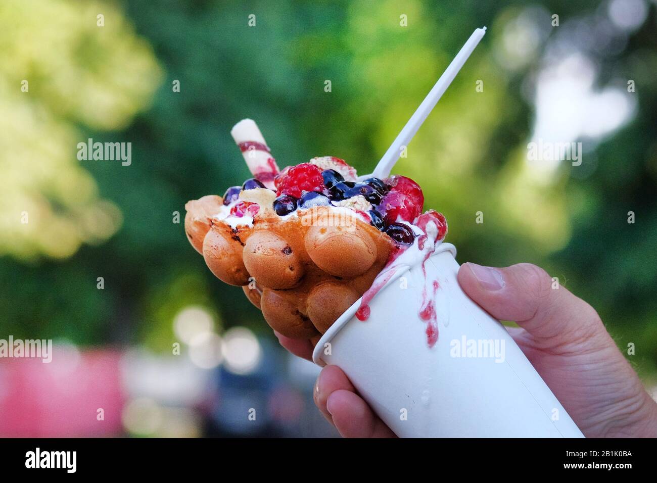 Berry nut bubble waffle in white paper cup in hand on blurred background. Stock Photo