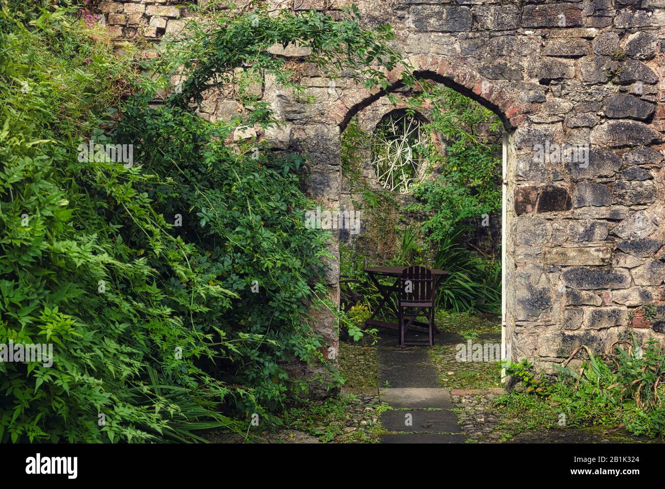 Rustic old stone archway in an overgrown secluded garden with climbing plants  and tables and chairs Stock Photo