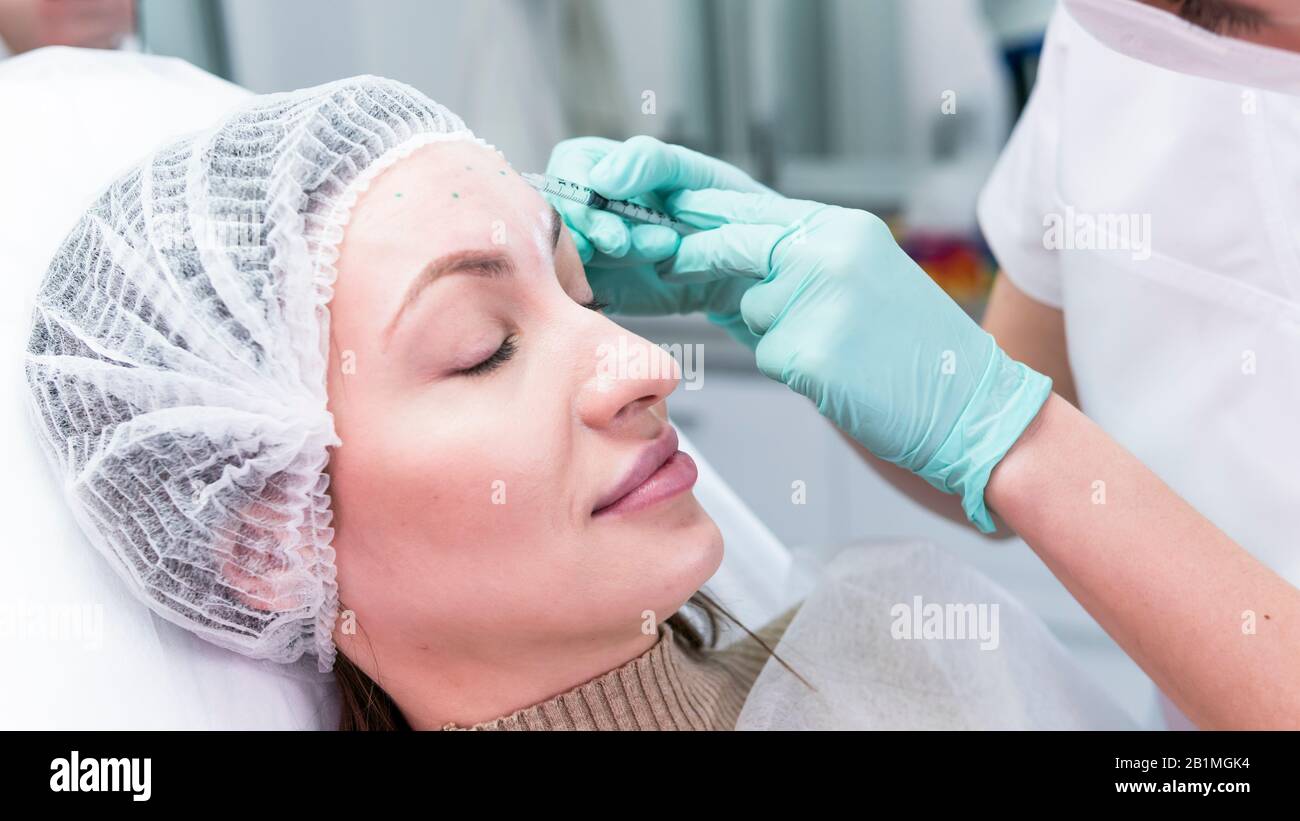 The young beautician doctor preparing to making injection in female forehead. The doctor cosmetologist makes anti-aging treatment and face lift proced Stock Photo