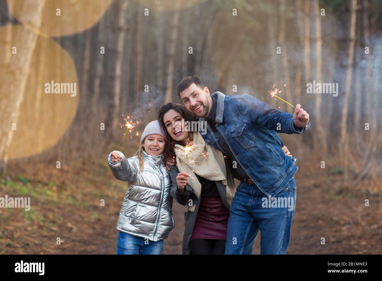 Cheerful young family with daughter dressed in warm clothes with sparklers in hands embracing and looking at camera while standing against forest back Stock Photo