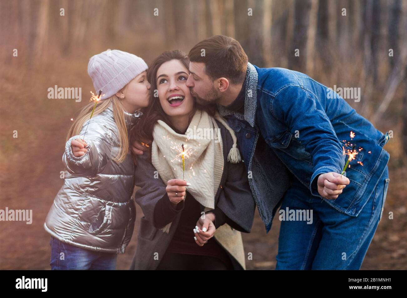Cheerful young family with daughter dressed in warm clothes with sparklers in hands embracing and looking at camera while standing against forest back Stock Photo