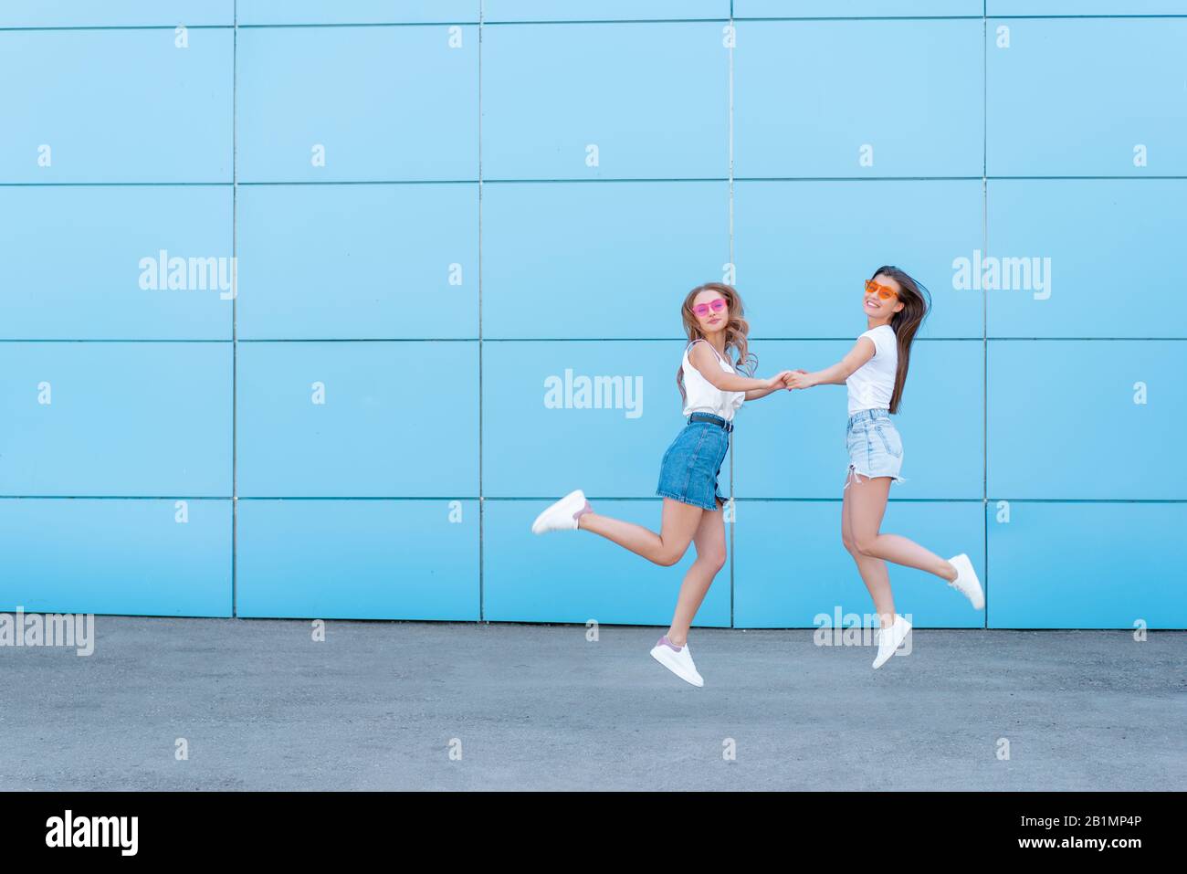two young hipster woman friends in retro neon sunglasses standing and smiling over blue wall Stock Photo