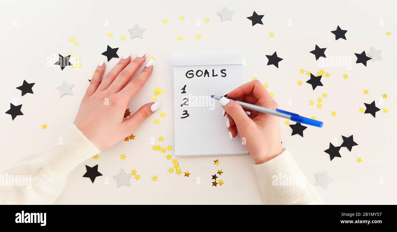 Female hand with pen writing New Year goals in empty notebook on white background with confetti Stock Photo