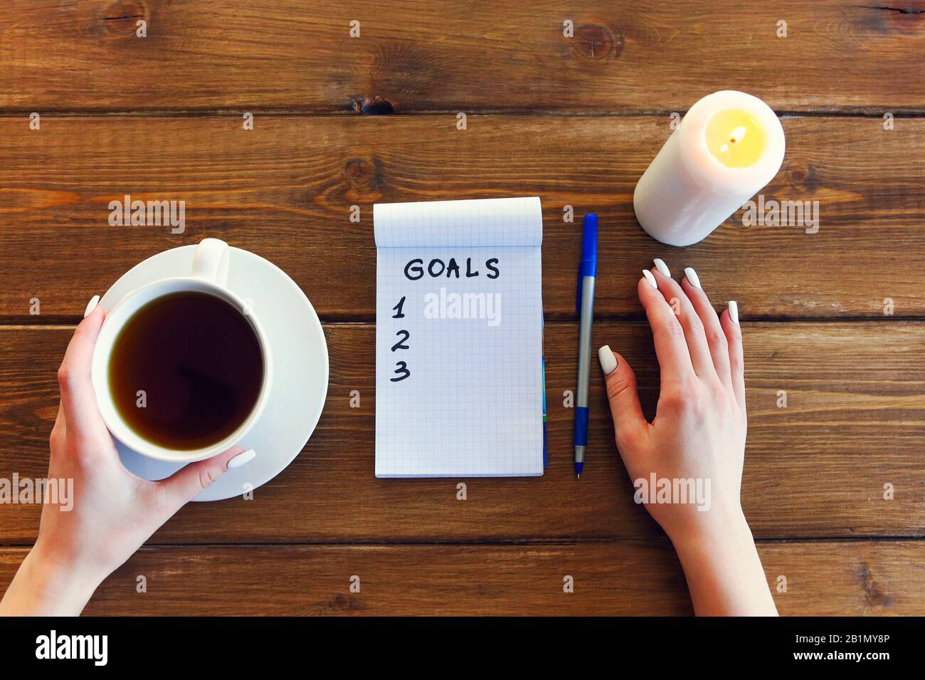 Female hand with pen writing goals in empty notebook on wooden table background with coffee and burning candle Stock Photo