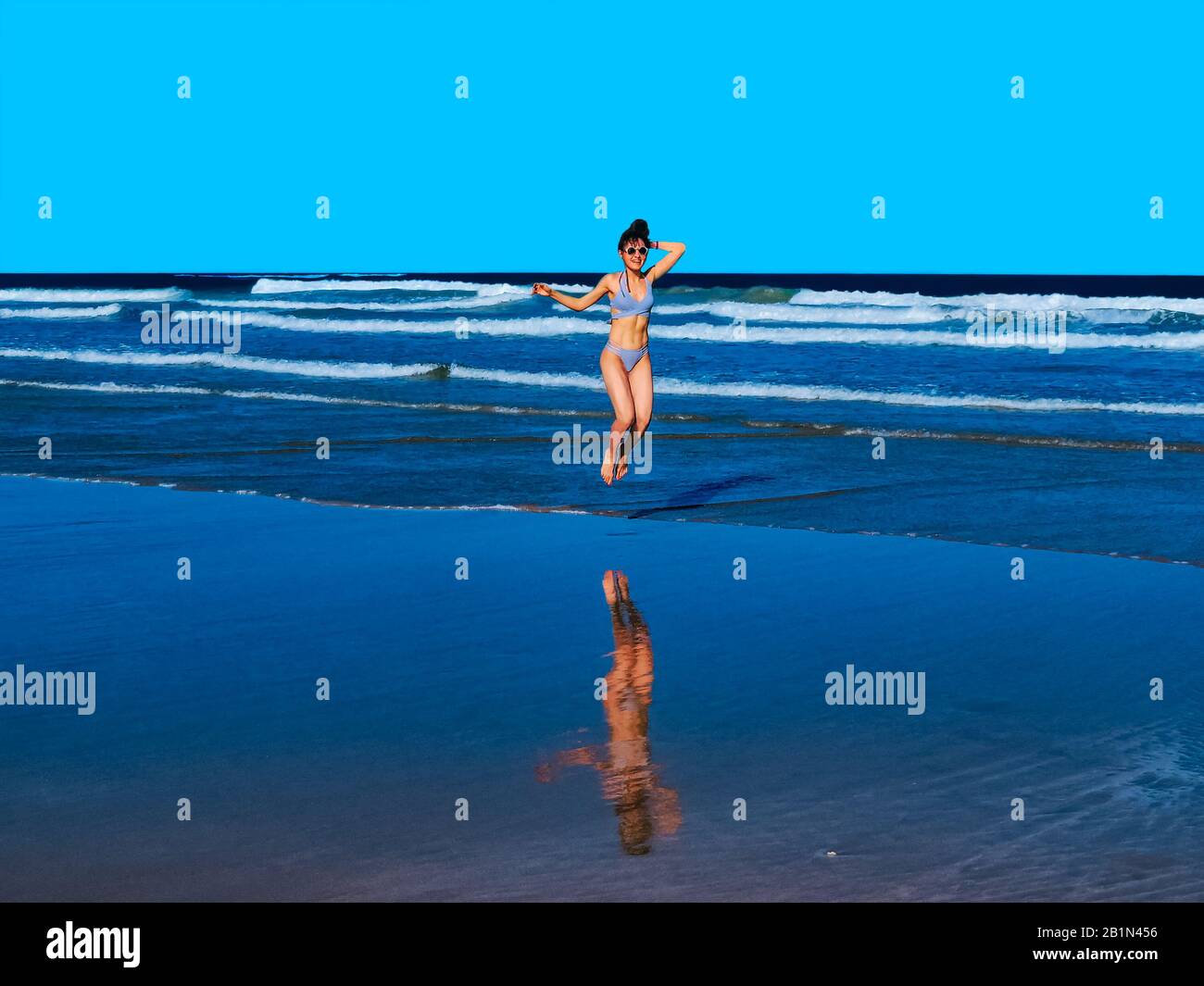 Happy young woman with a beautiful body wearing bikinis showing abs and jumping on beach shore - Blue highlights and shadows - Summer Trip Concept Stock Photo