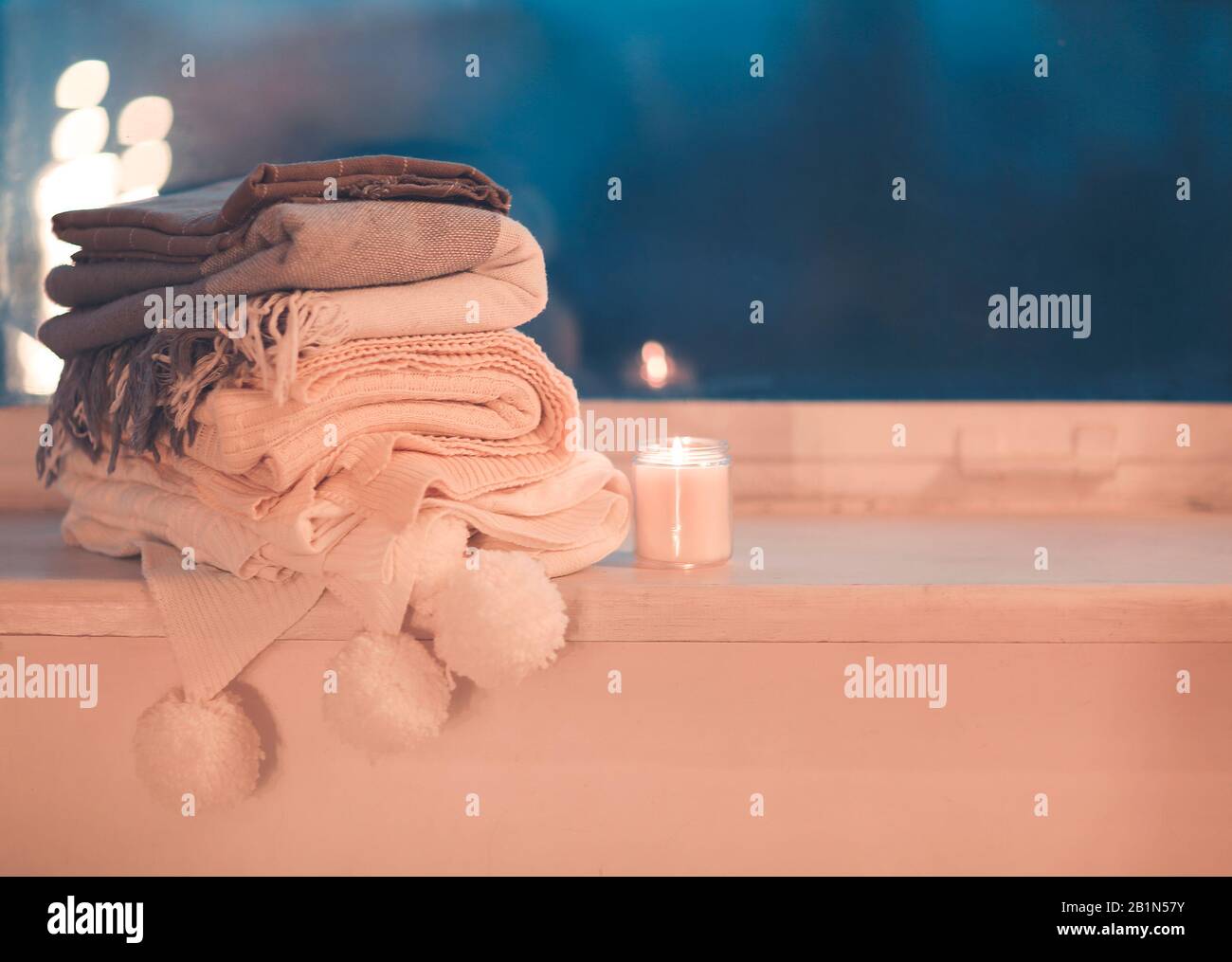 Stack of knitted plaids with gray mug and burning candle on cozy window Stock Photo