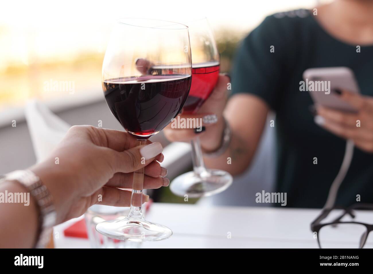 two hands of a girl in a beautiful restaurant at a table holding glasses with a red drink of champagne wine at dinner, raising glasses during a toast Stock Photo