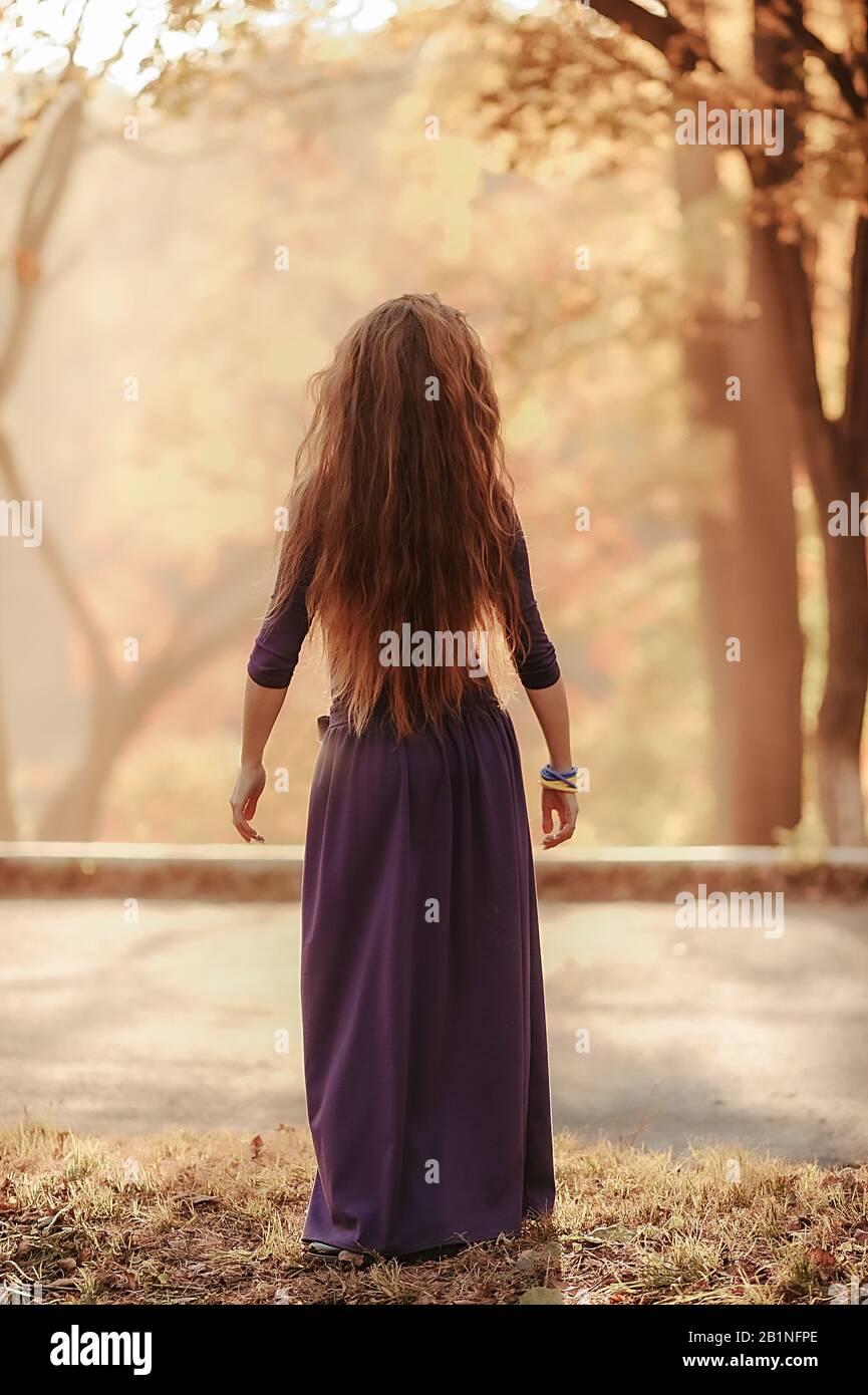 a sophisticated slender girl with long curly hair in a purple floor-length dress dances an Oriental dance against the background of the autumn forest Stock Photo