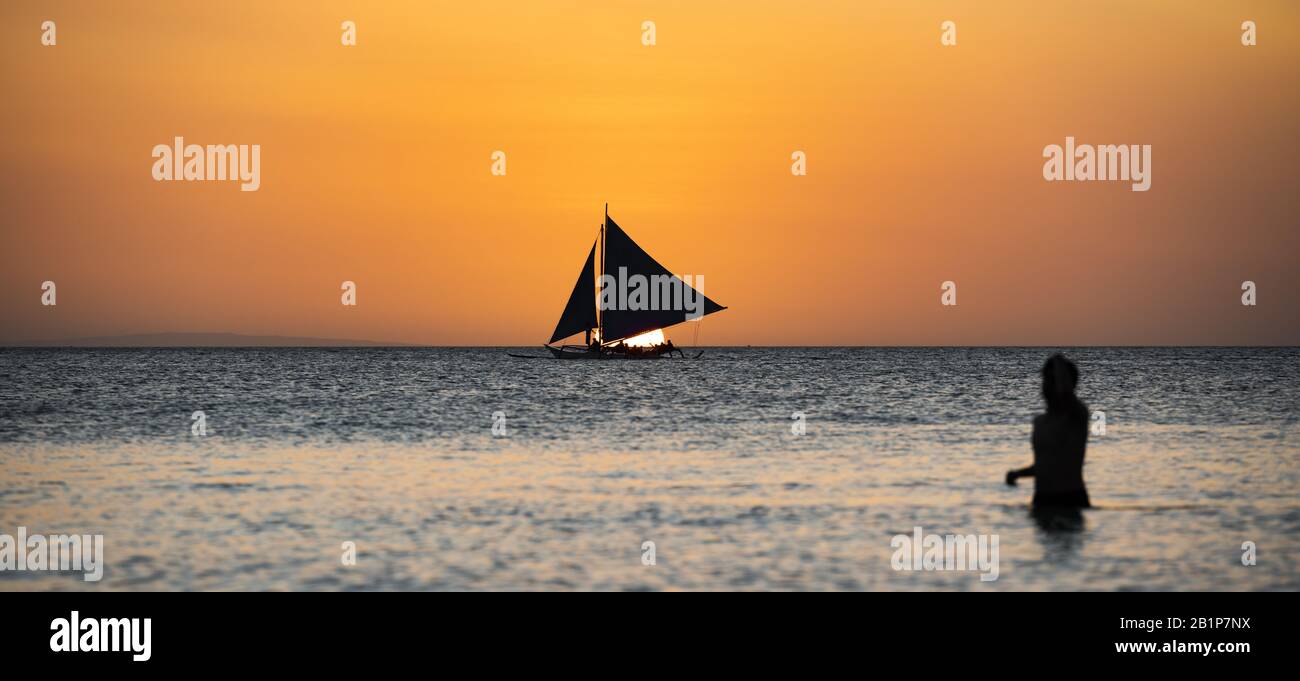 (Selective focus) Stunning view of a boat sailing during a beautiful sunset in the background and the silhouette of a blurred person in the foreground. Stock Photo